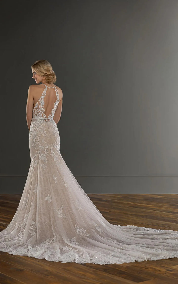 A woman wearing a bridal gown is shown from the back in a studio setting with a dark gray wall and wooden floor. The dress has a fitted lace mermaid silhouette with a long train and an open back.