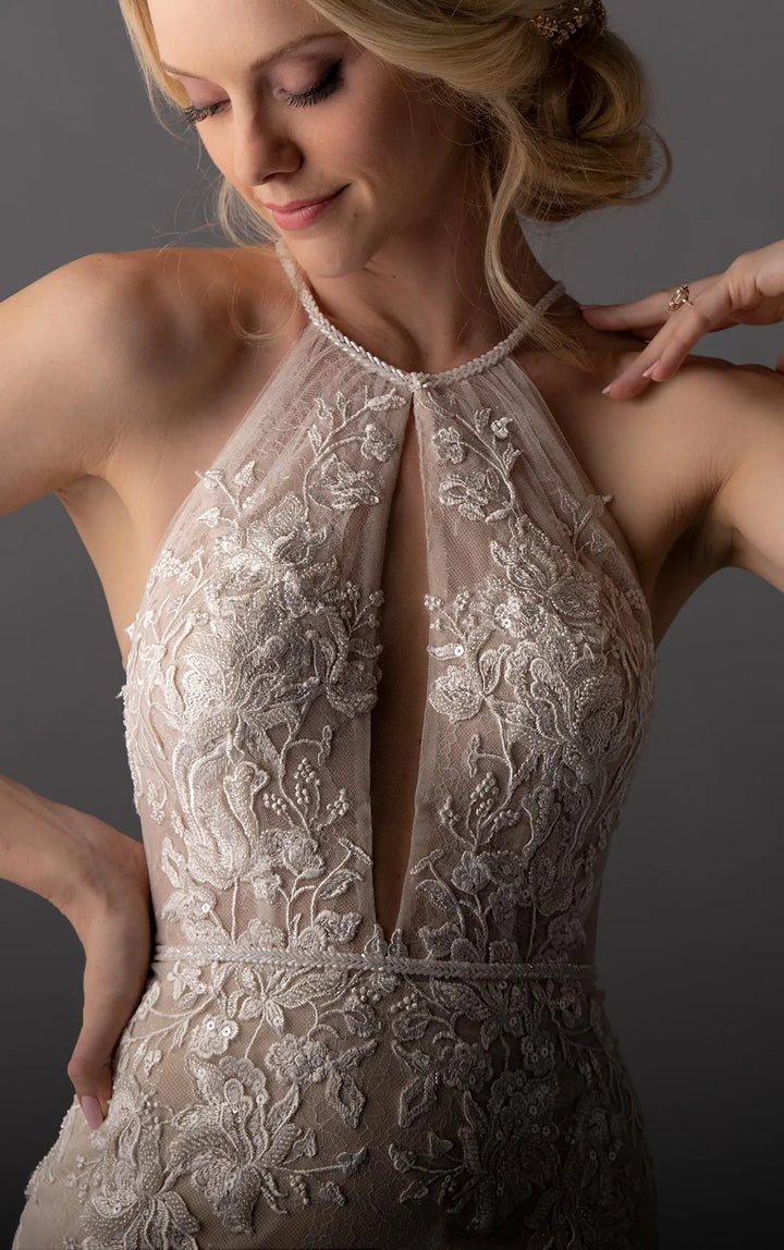 A close-up of a woman wearing a sleeveless halter-neck dress with intricate floral lace and bead embroidery in a champagne beige tone against a dark gray studio background.