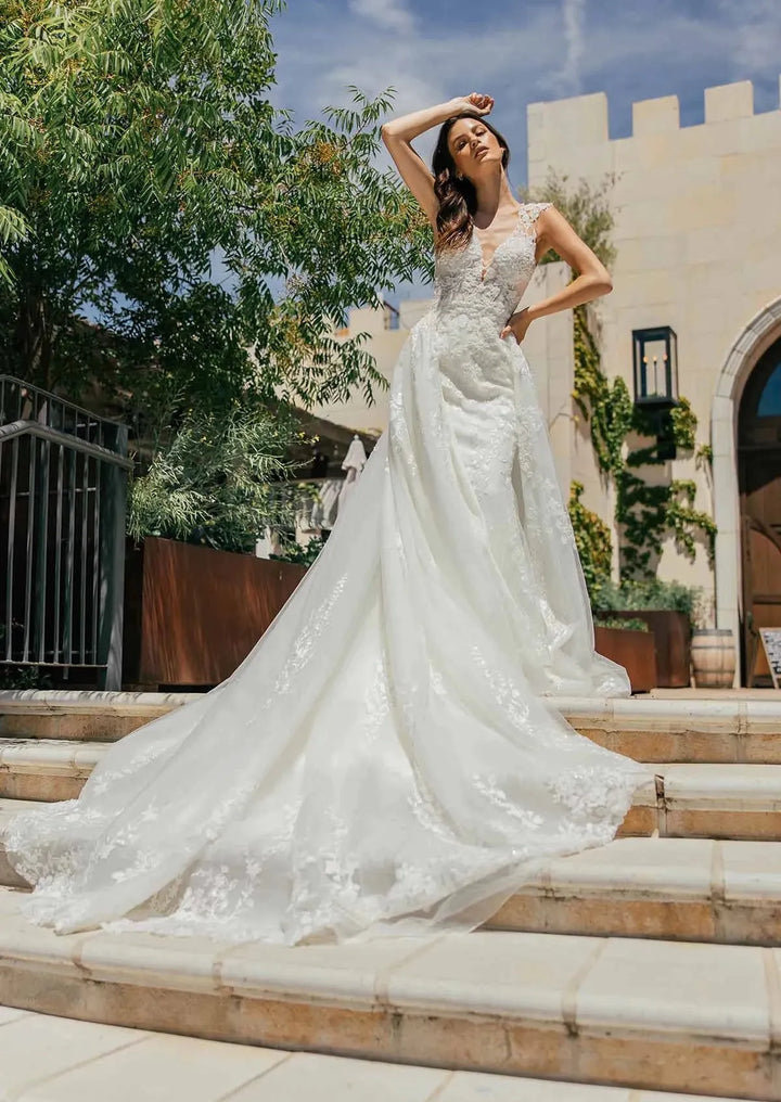 A woman wearing a sleeveless white lace wedding gown with a long train stands on outdoor stone steps in front of a beige building under a blue sky.