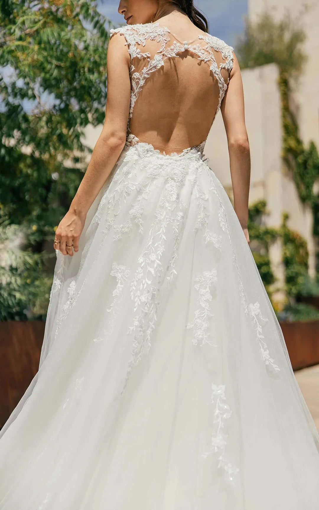 A woman wearing a white lace wedding gown shown from the back, featuring a sheer open back and a full tulle skirt, photographed outdoors with blurred greenery and a building in the background.