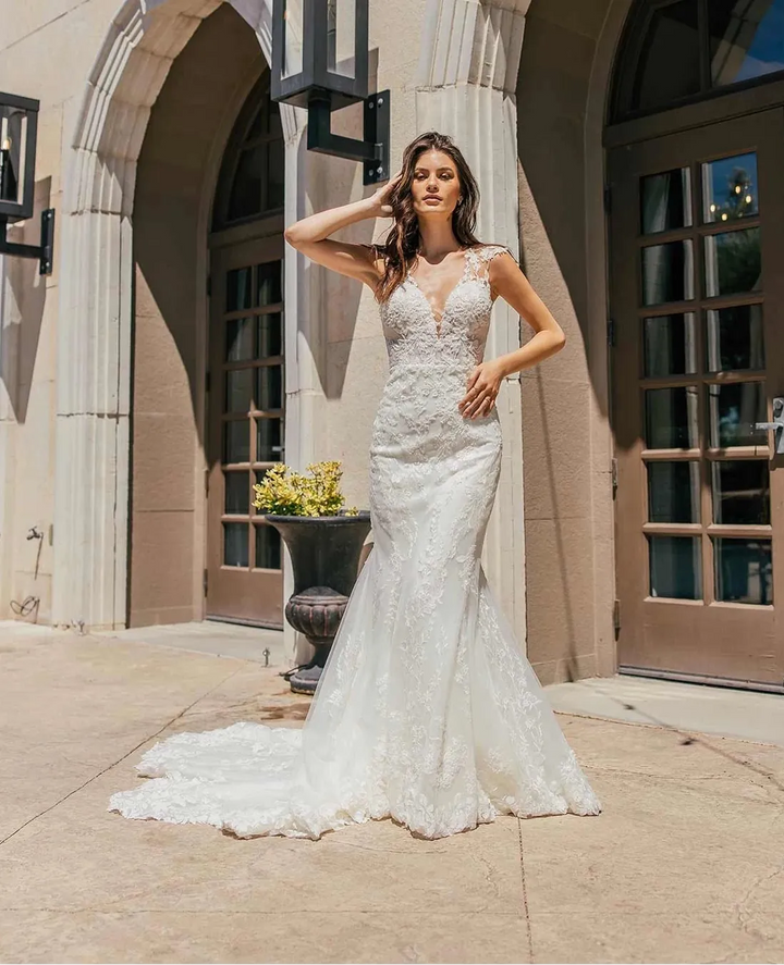 A woman wearing a white lace mermaid-style wedding gown stands outdoors in front of a beige stone building with arched doorways and windows.