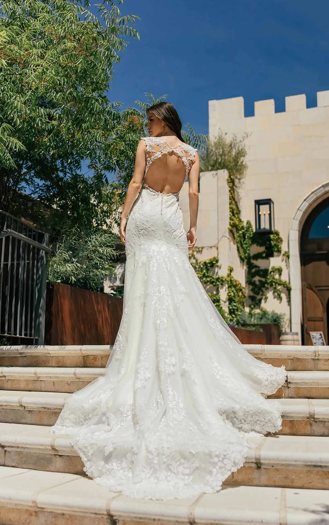 A woman wearing a white lace mermaid-style wedding gown with an open back and short sleeves, standing on outdoor stone steps in front of a beige building with greenery and blue sky.