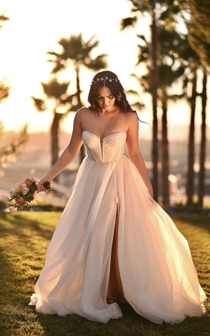 A woman wearing a strapless sweetheart wedding gown with a fitted corset-style bodice and a flowing tulle skirt, standing outdoors on grass with palm trees and warm sunset light in the background.