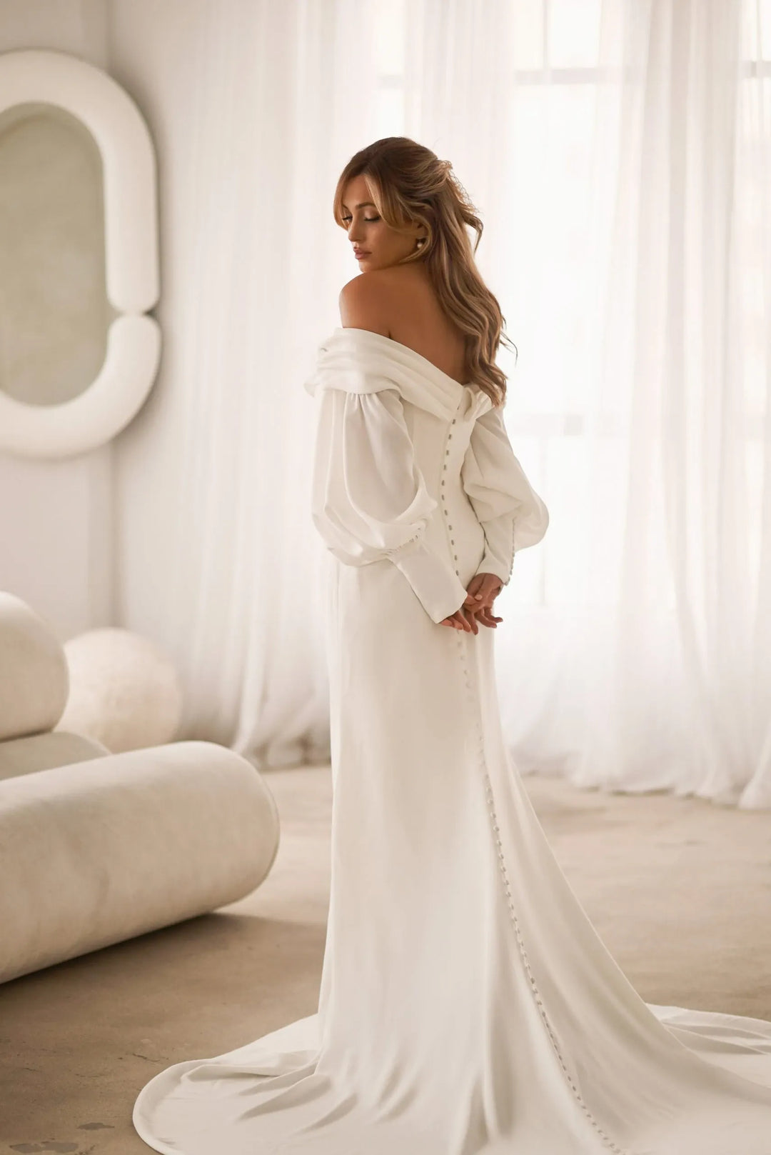 A woman wearing an off-the-shoulder white wedding gown with long balloon sleeves and a train, standing in a bright, softly lit room with sheer curtains and a sofa.