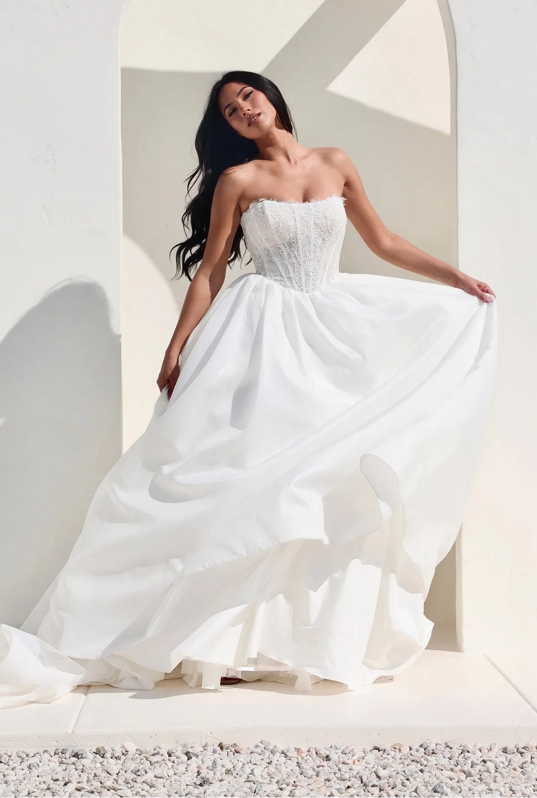 A woman modeling a strapless white wedding gown with a structured bodice and full flowing skirt, posed in front of a white arched architectural setting with strong sunlight and shadows.
