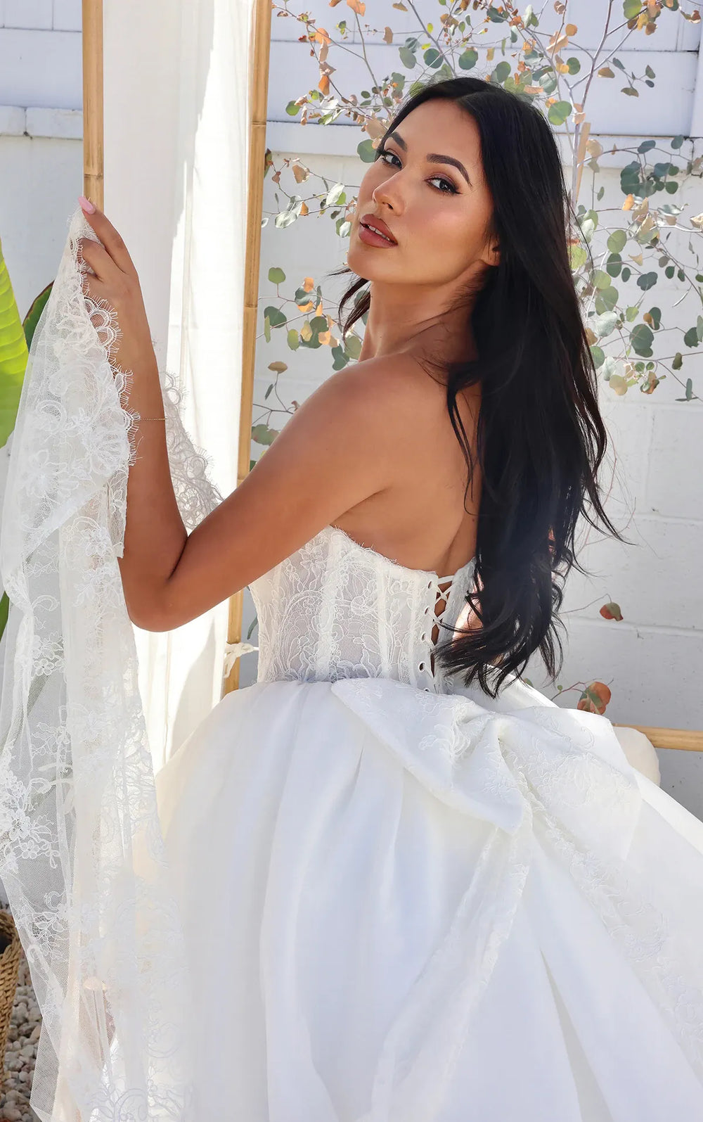 A woman wearing a strapless white wedding gown with lace detailing and a voluminous skirt, posed in front of a white wall with leafy branches.