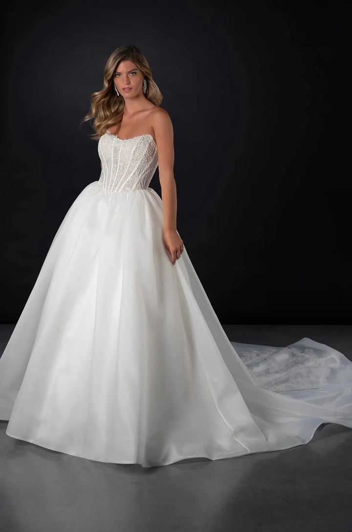 A woman modeling a strapless white ball gown wedding dress with a fitted lace bodice and a full tulle skirt with a long train, posed against a dark studio background.
