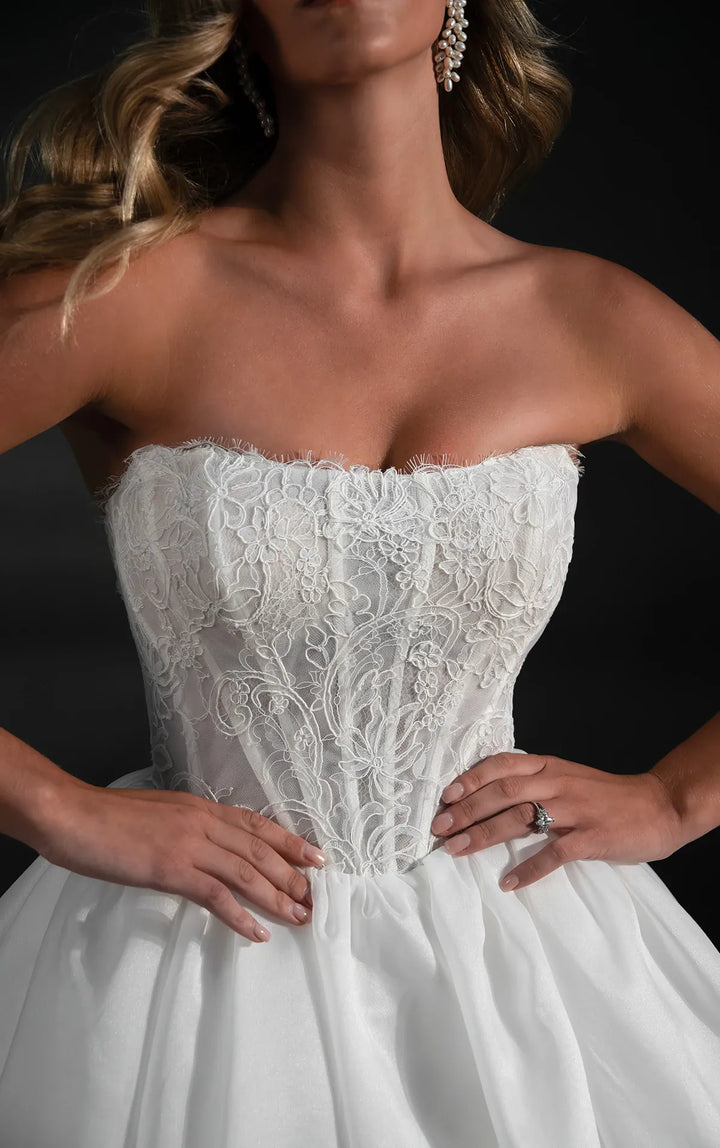 A close-up of a model wearing a strapless white lace wedding gown with a fitted lace bodice and a full tulle skirt against a dark background.