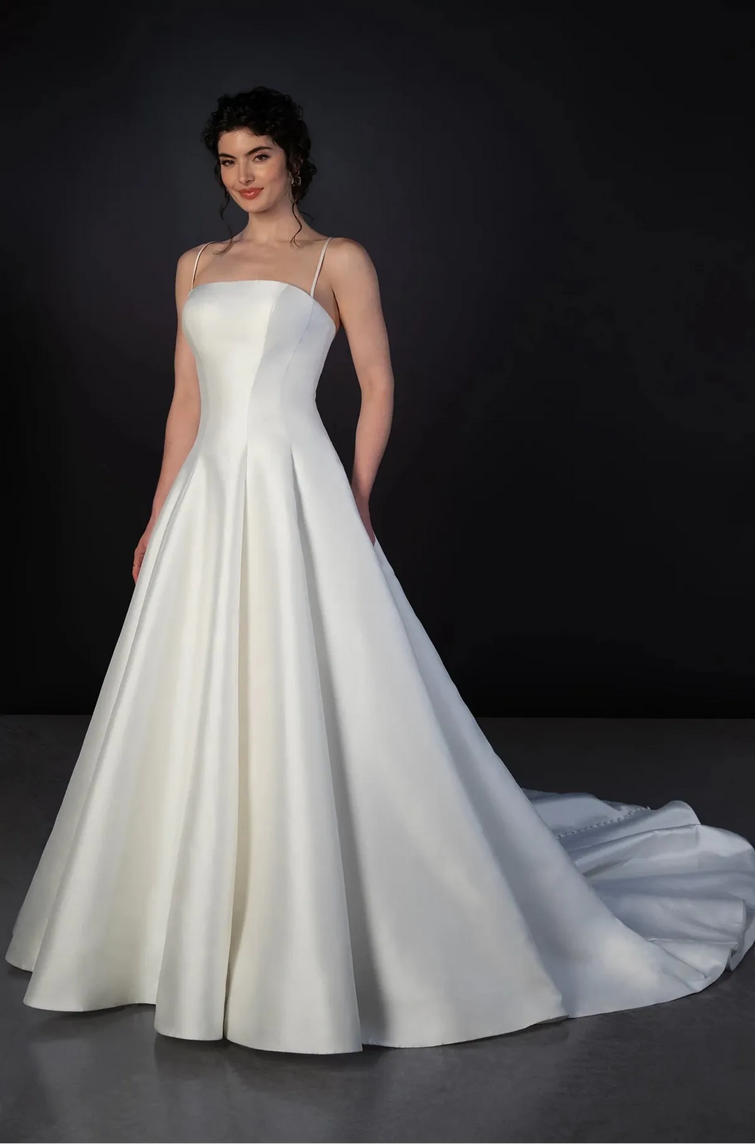 A woman modeling a white strapless A-line wedding gown with a fitted bodice, full skirt, and long train against a dark studio background.