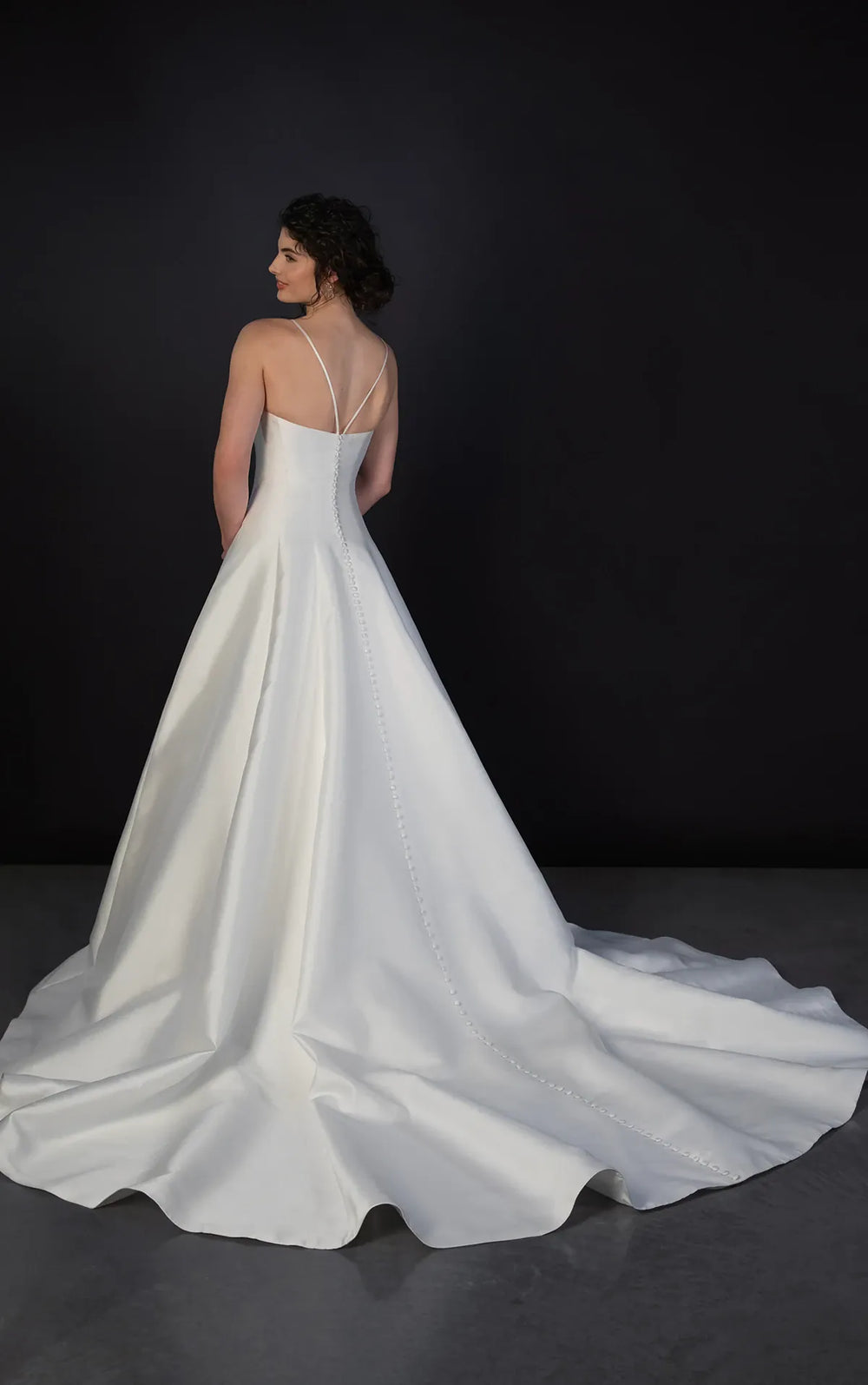 A woman wearing a white satin wedding gown with thin straps and a long train, photographed from the back against a dark studio background.