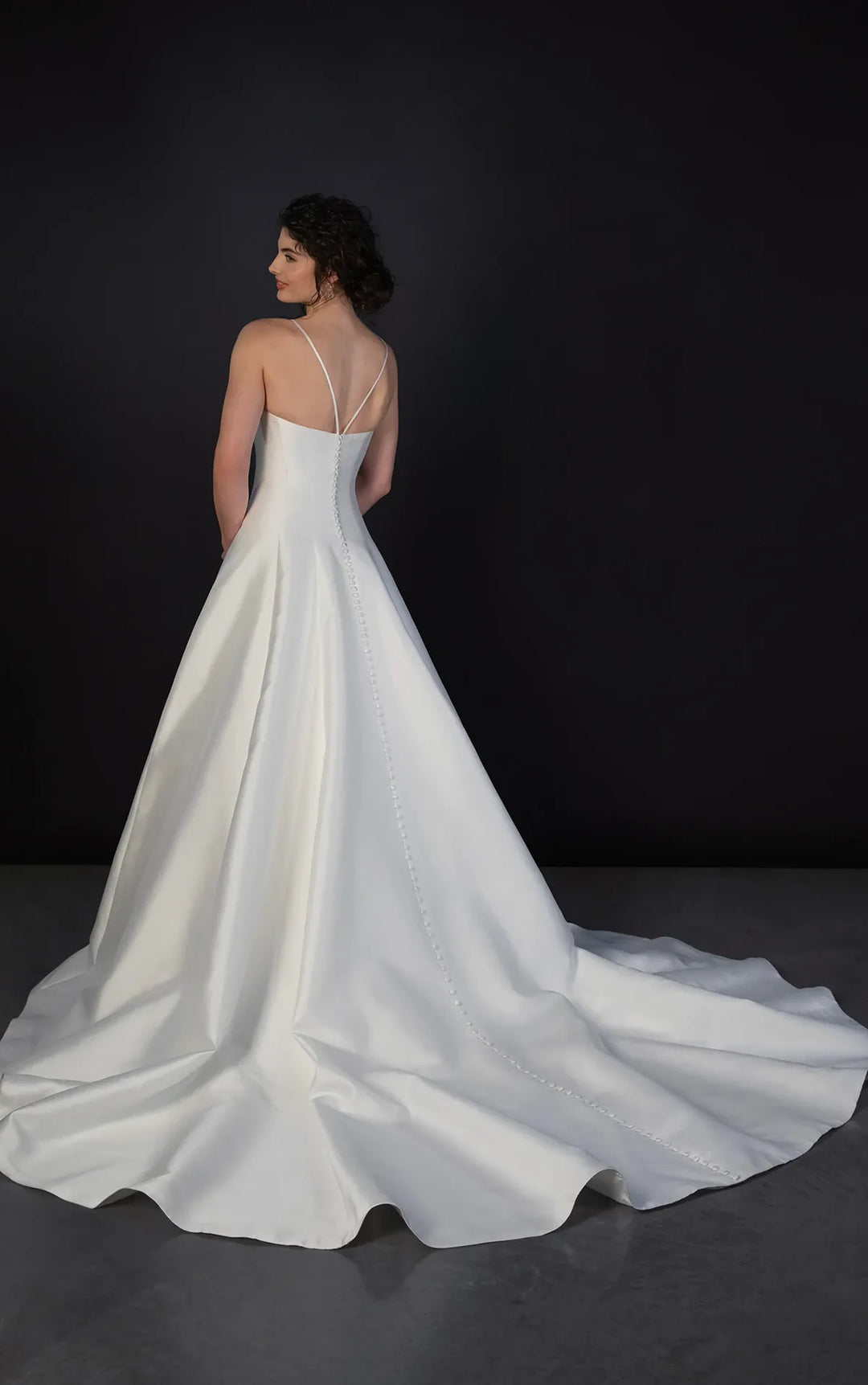 A woman wearing a white satin wedding gown with thin straps and a long train, photographed from the back against a dark studio background.