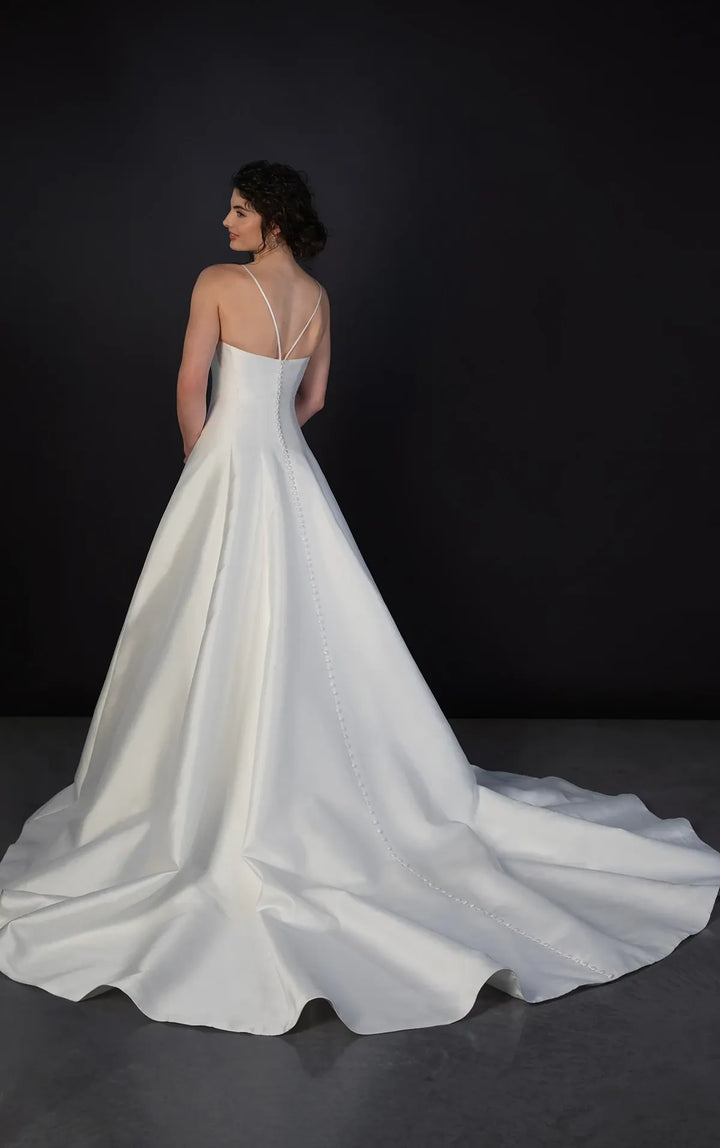 A woman wearing a white satin wedding gown with thin straps and a long train, photographed from the back against a dark studio background.