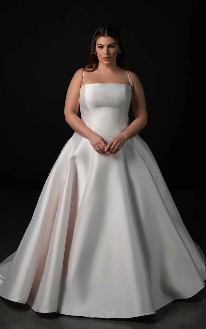 A woman modeling a white ball gown wedding dress with a square neckline, thin straps, and a full satin skirt against a dark studio background.