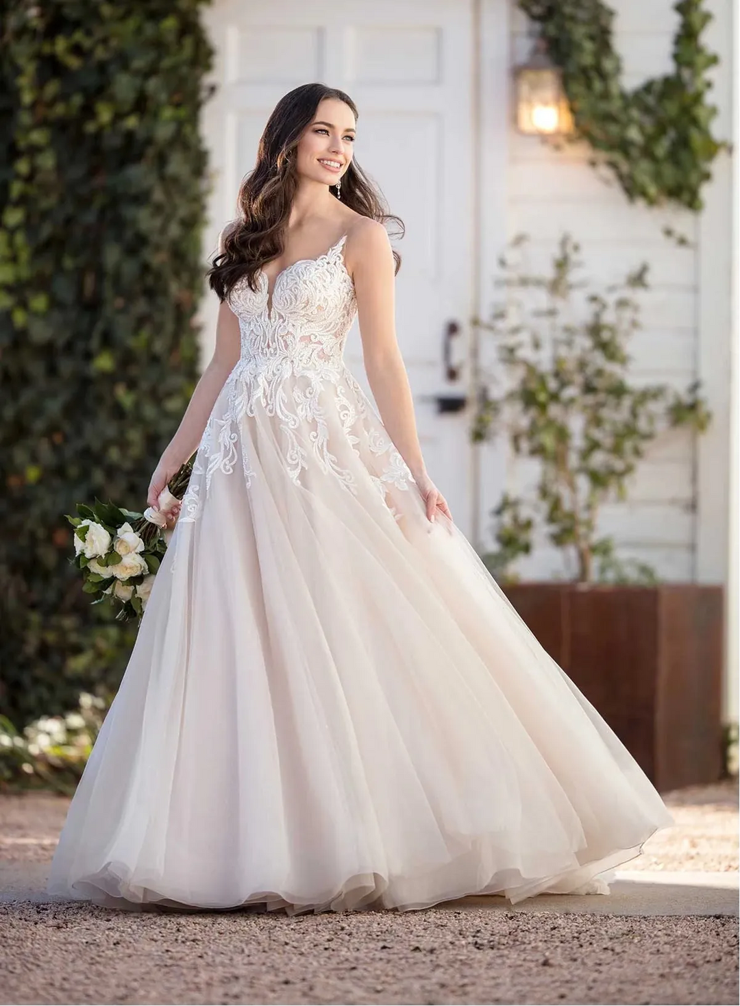 Woman in a white wedding dress standing outdoors with greenery and a white door in the background.
