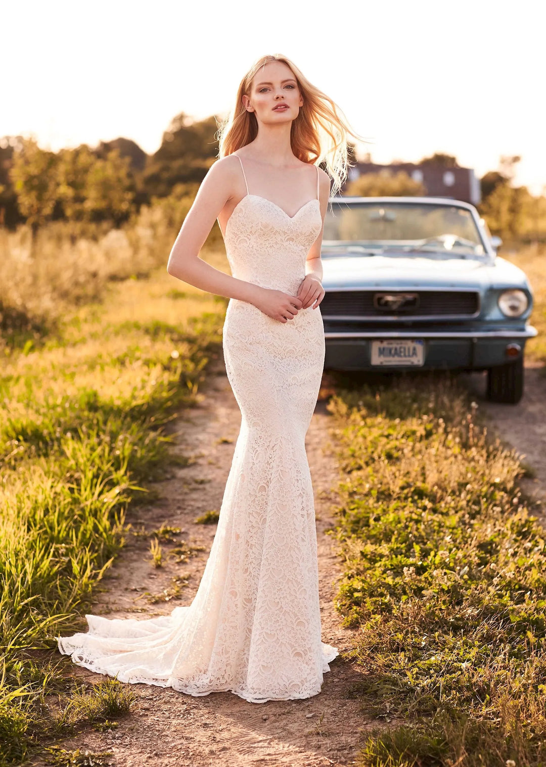 Woman in a white lace dress standing on a dirt road with a vintage car in the background.