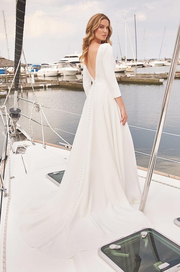 A woman wearing a white wedding gown stands on a sailboat deck with a marina and boats in the background. The dress has three-quarter sleeves, a deep V-back, and a long train.