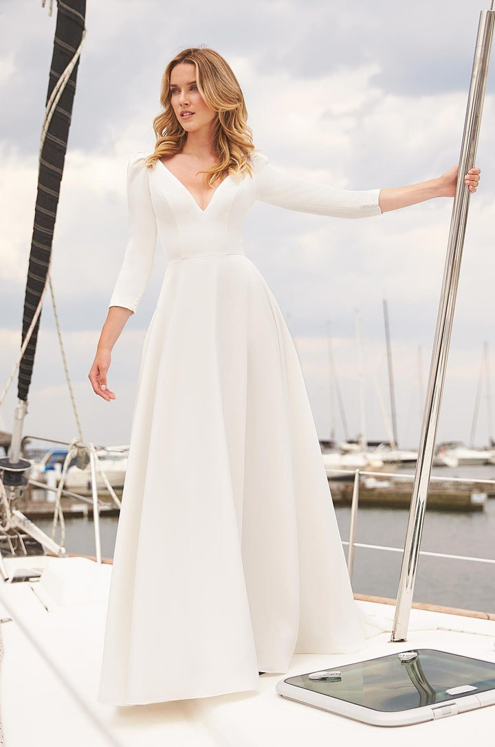 A woman wearing a long-sleeved white A-line wedding dress stands on a sailboat deck with a marina background.