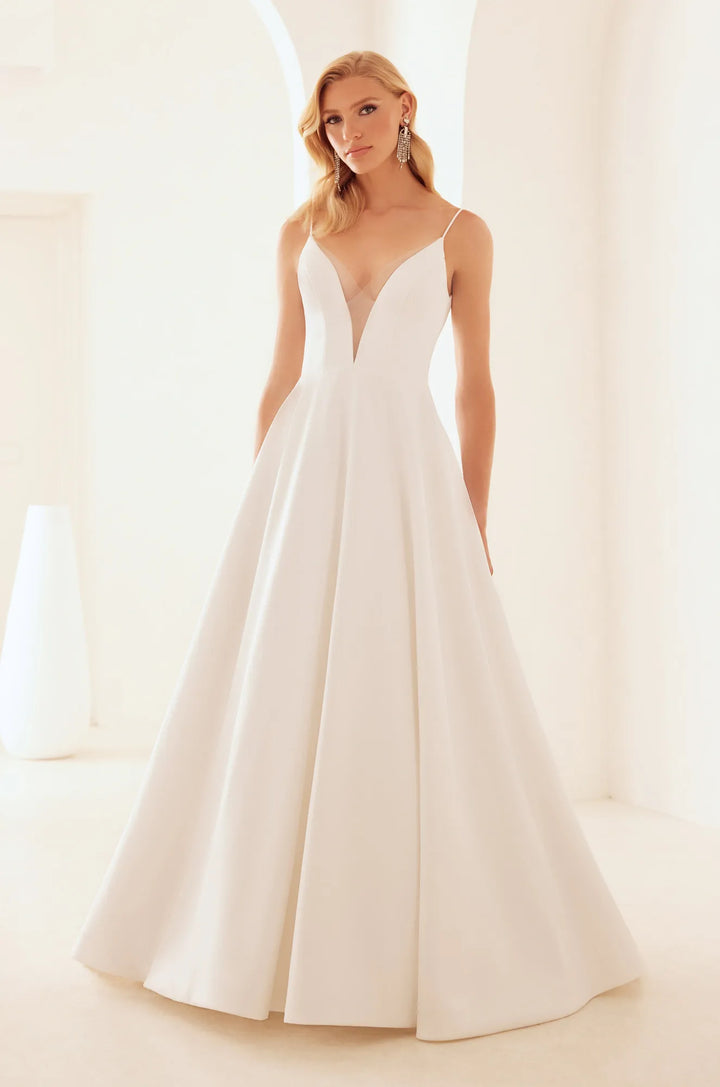 A woman modeling a white A-line wedding dress with thin spaghetti straps, a deep V-neckline, and a full skirt in a bright, minimalist white interior.