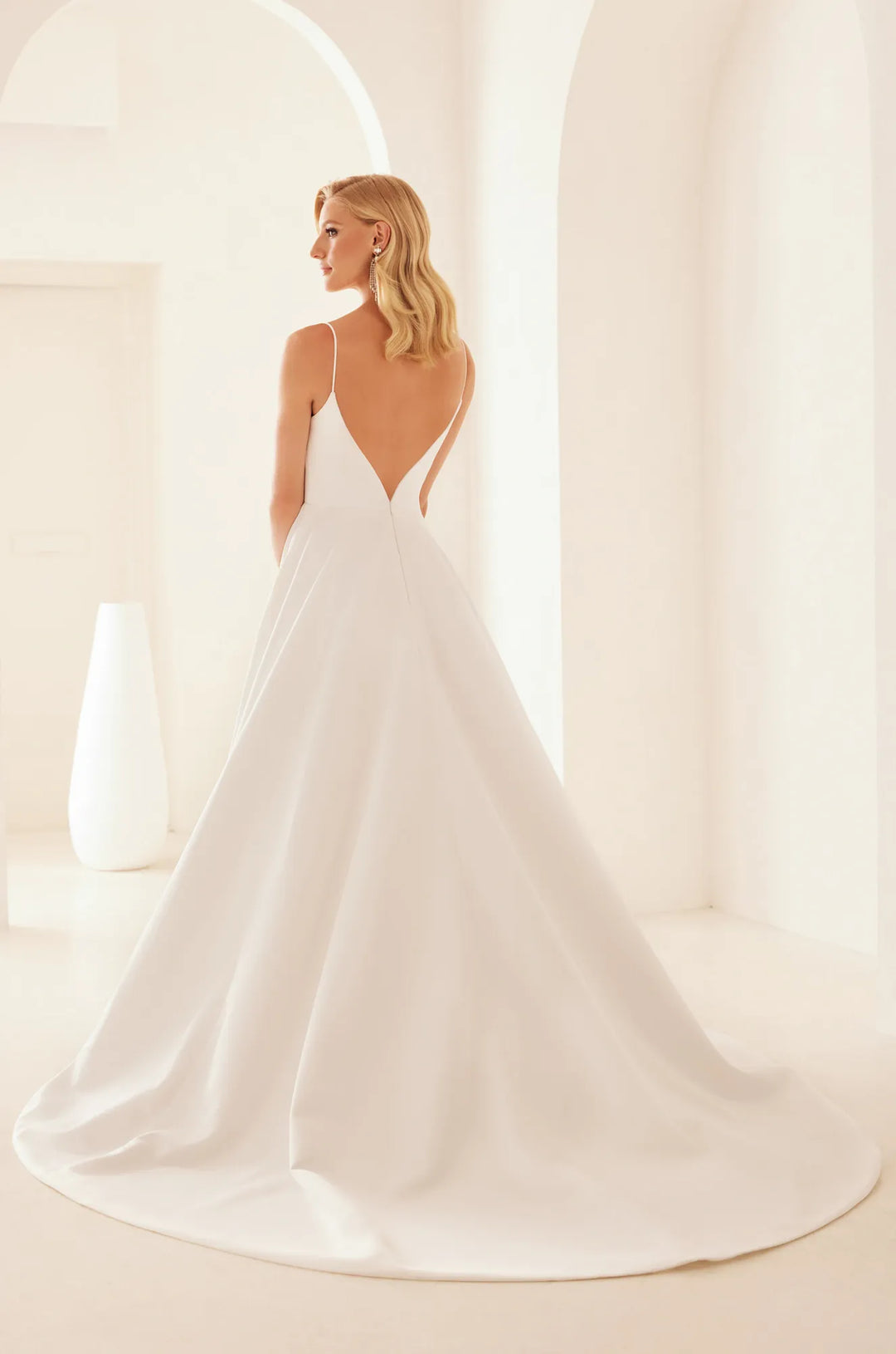 A woman wearing a white bridal gown with thin straps, a deep V back, and a full skirt with a long train, photographed in a bright, minimalist interior with arches.
