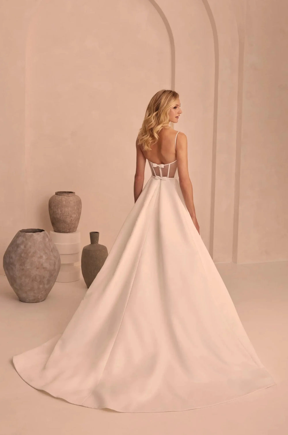 A woman wearing a voluminous white wedding gown with a low open back and sheer upper bodice, photographed in a softly lit beige interior with arches and decorative vases.
