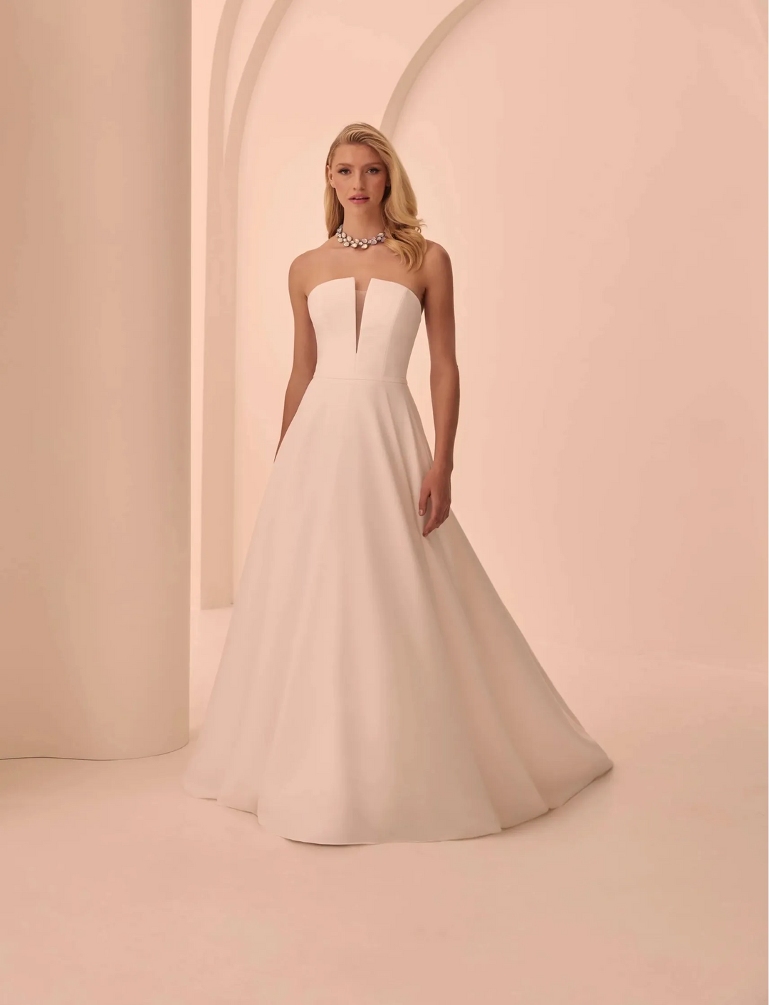 A woman wearing a strapless white wedding gown with a fitted bodice and full skirt, standing in a minimalist cream-colored studio with arches and columns.