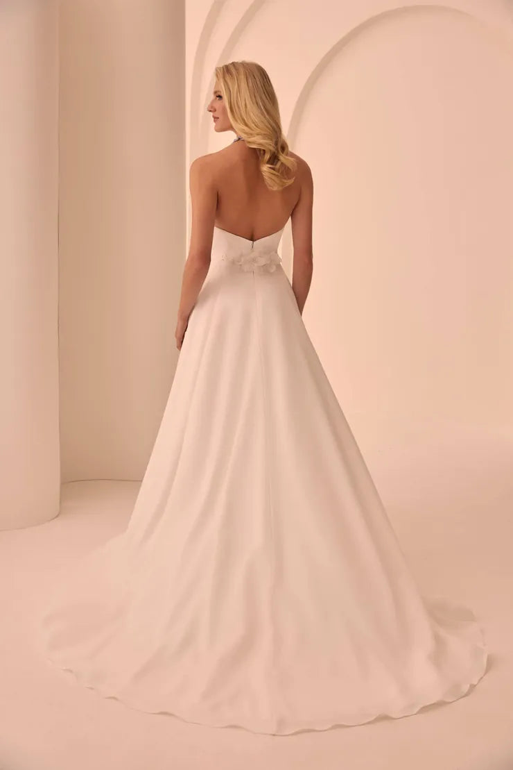 A woman modeling a strapless A-line wedding gown from the back, showing a low open back, floral applique at the waist, and a full flowing skirt in a soft blush-ivory tone against a warm cream studio backdrop.