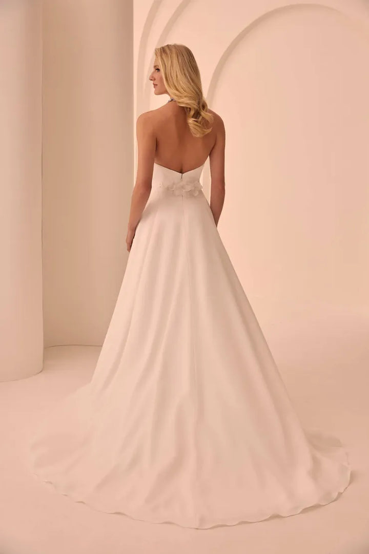 A woman modeling a strapless A-line wedding gown from the back, showing a low open back, floral applique at the waist, and a full flowing skirt in a soft blush-ivory tone against a warm cream studio backdrop.