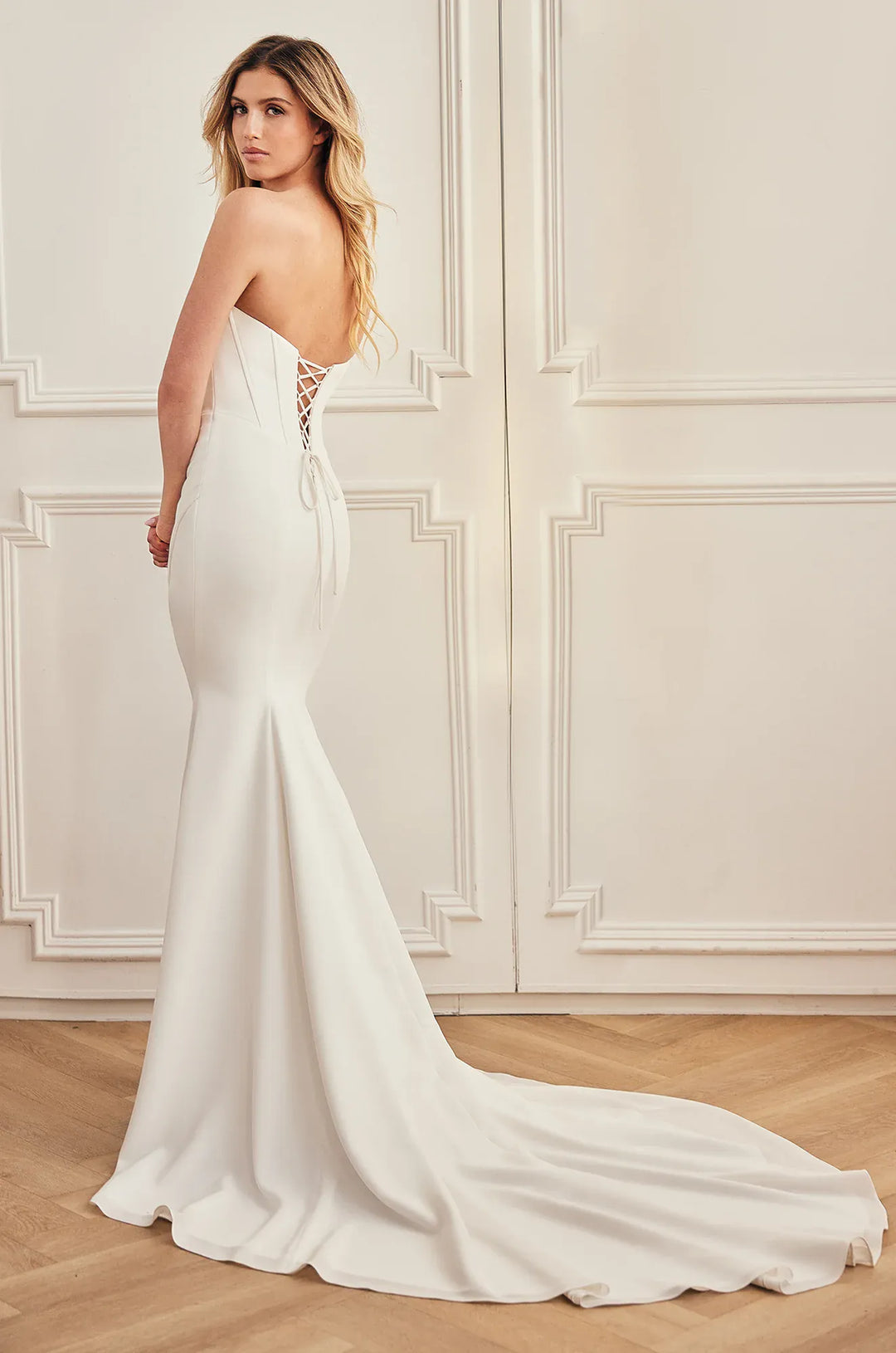 A woman modeling a strapless white mermaid-style wedding gown with a lace-up open back, shown from the rear in a bright indoor room with cream paneled walls and wood flooring.