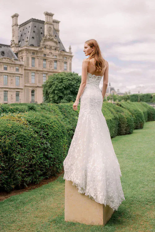 A woman wearing a strapless lace wedding gown stands on a stone pedestal in a garden, with a large historic building in the background.
