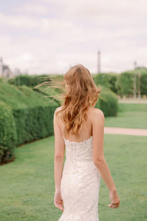 A woman with long wavy light brown hair seen from behind, wearing a strapless white lace wedding dress and walking on a green lawn in an outdoor garden setting.