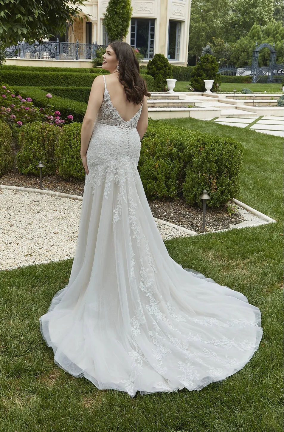 Woman in a white lace wedding dress standing on grass with a building and garden in the background