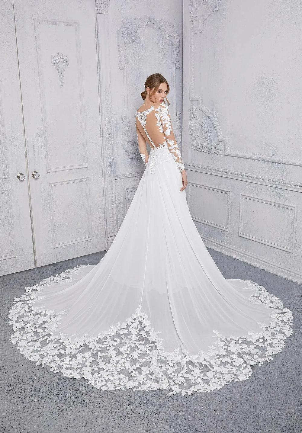 Woman wearing a white wedding dress with floral lace details in a room with gray walls.