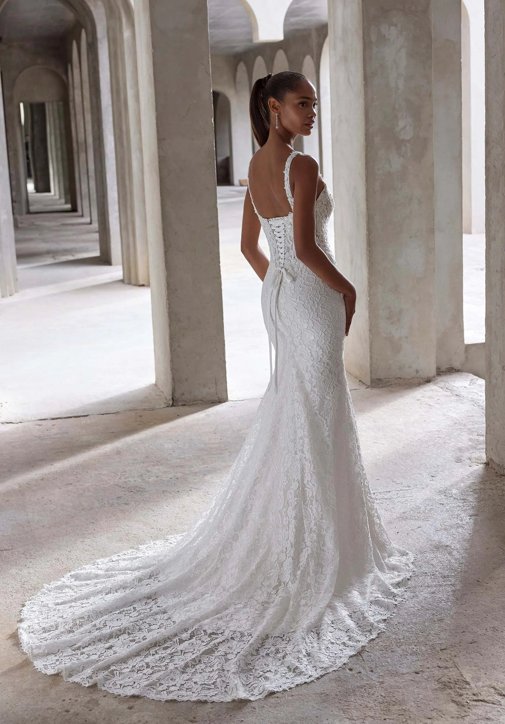 Woman in a white lace wedding dress standing in a sunlit hallway with columns.