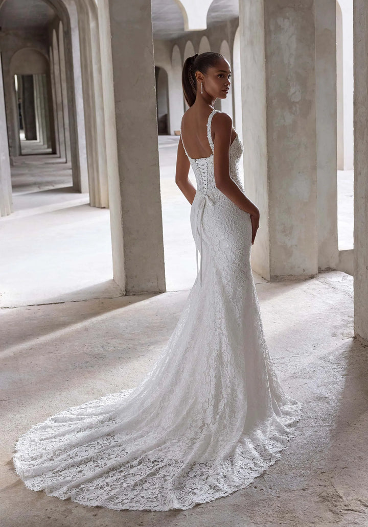 Woman in a white lace wedding dress standing in a sunlit hallway with columns.