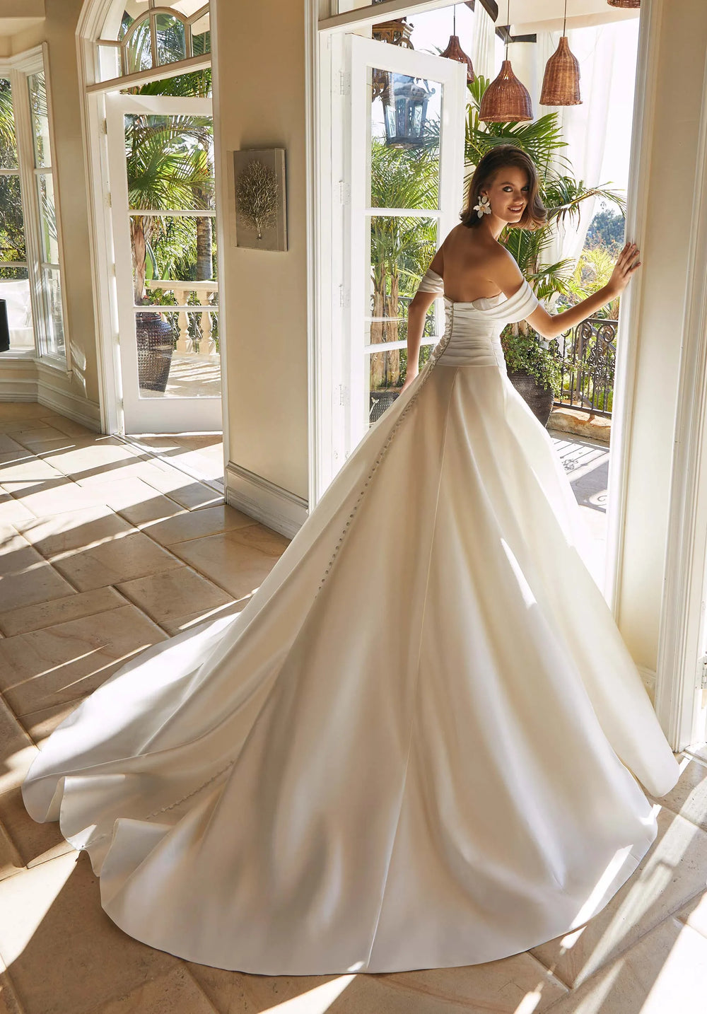 Woman in a white wedding dress standing in a sunlit room with large windows.