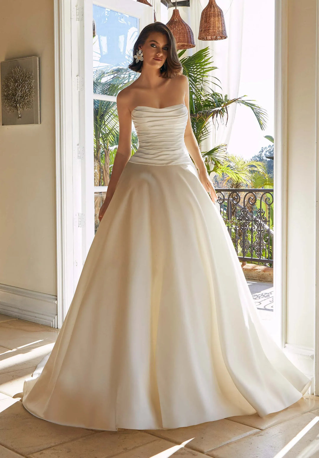 A woman models a strapless ball gown wedding dress with a ruched bodice and a full, flowing skirt, standing in a sunlit interior doorway with plants visible outside.