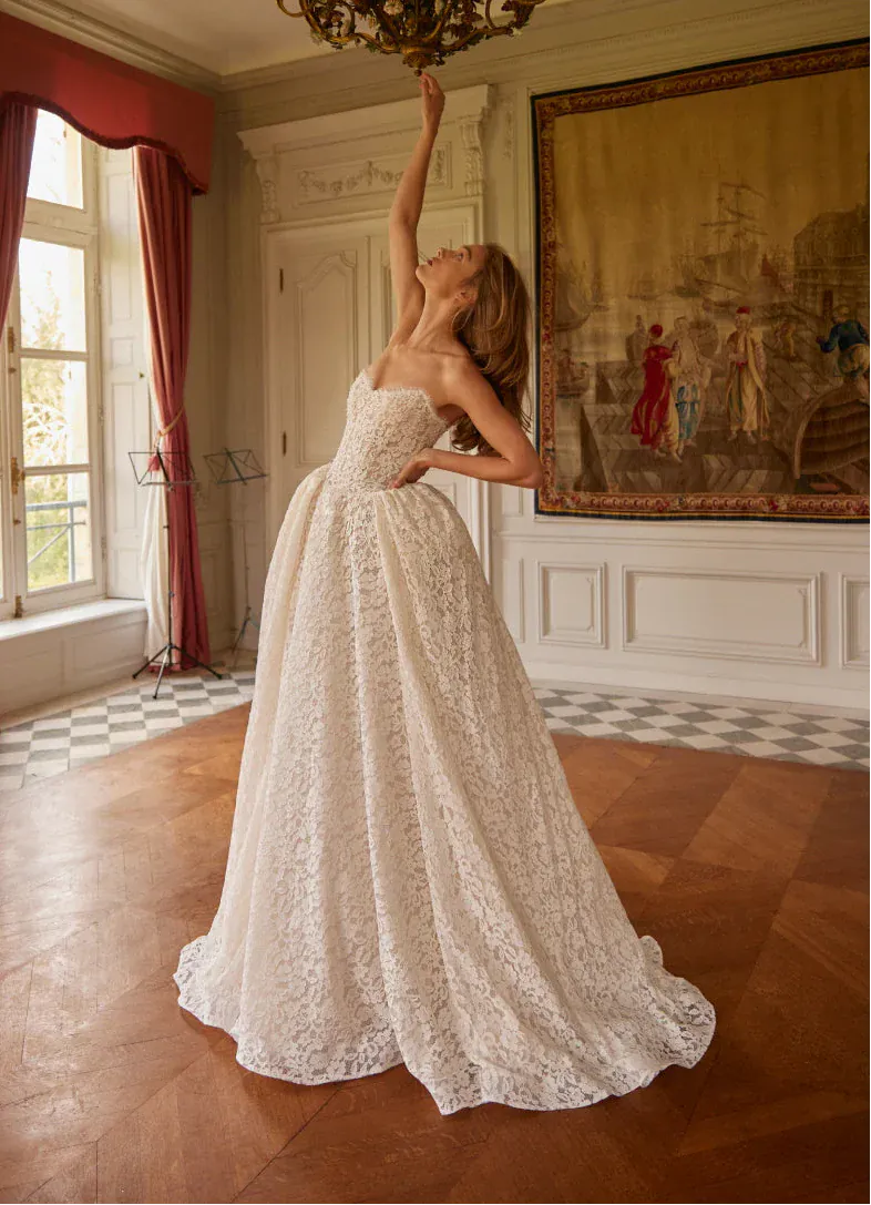Woman in a white lace wedding dress reaching towards a chandelier in an elegant room.