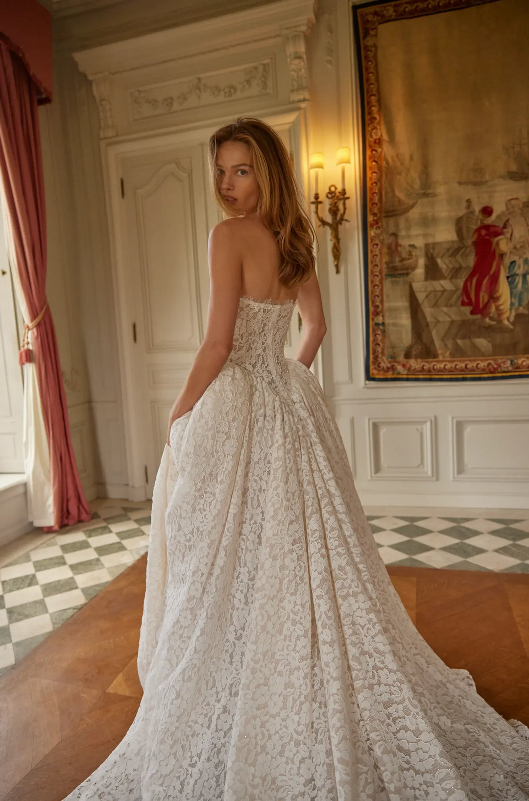 Woman in a lace wedding dress standing in an elegant room with decorative wall art.