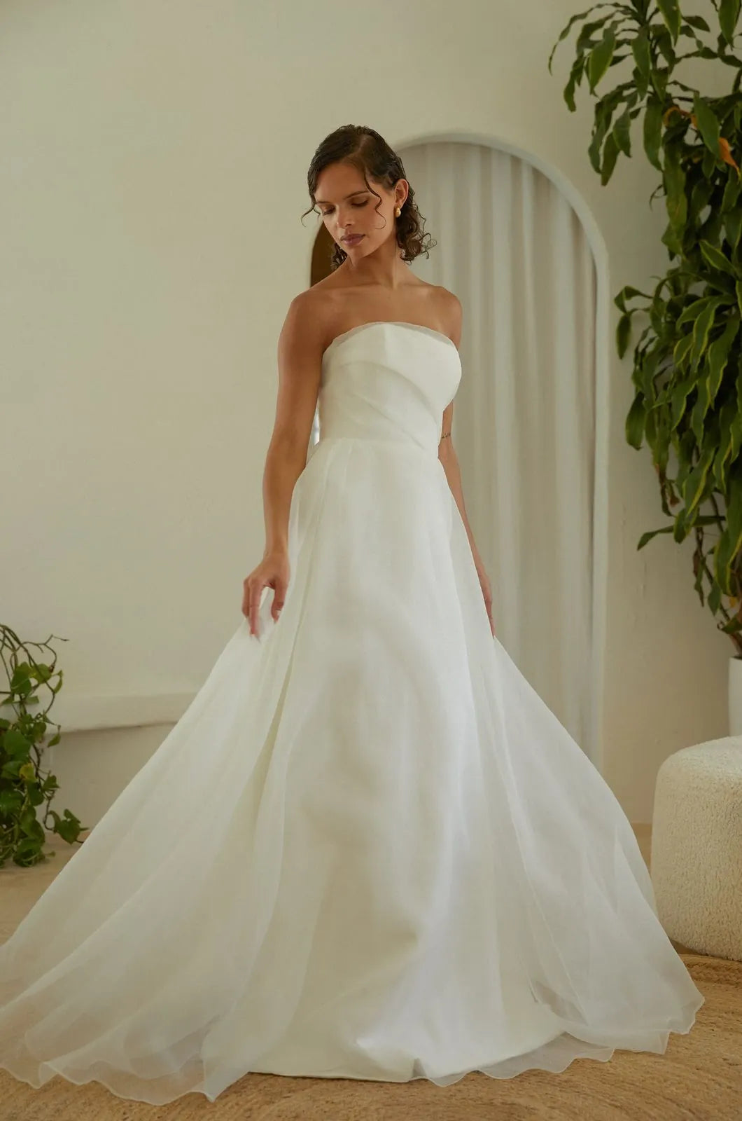 A woman wearing a strapless white A-line wedding gown with a structured bodice and flowing tulle skirt, standing indoors in a softly lit room with plants and a cream-colored arched doorway behind her.