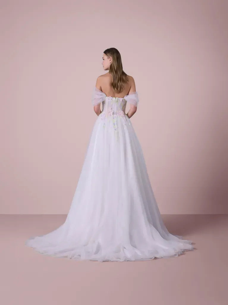 A woman stands with her back to the camera wearing a white off-the-shoulder ball gown with a sheer tulle skirt and delicate floral embroidery on the bodice, set against a solid dusty pink studio background.