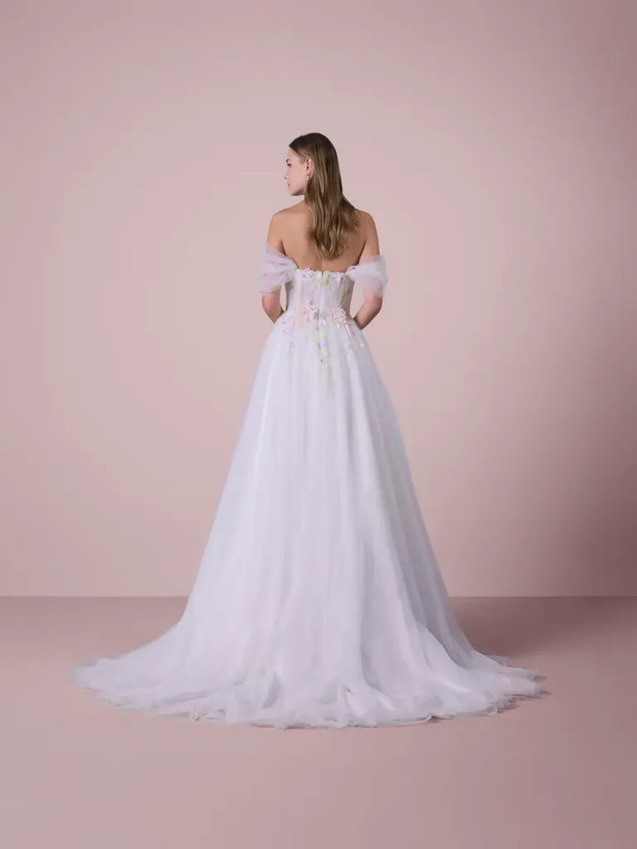 A woman stands with her back to the camera wearing a white off-the-shoulder ball gown with a sheer tulle skirt and delicate floral embroidery on the bodice, set against a solid dusty pink studio background.