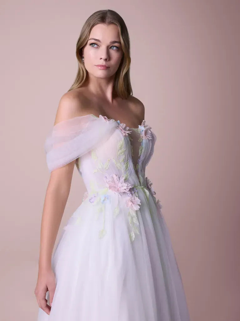 A woman modeling an off-the-shoulder white tulle gown with pastel pink and green floral embroidery, set against a solid pale pink studio background.