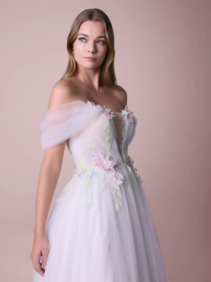 A woman modeling an off-the-shoulder white tulle gown with pastel pink and green floral embroidery, set against a solid pale pink studio background.