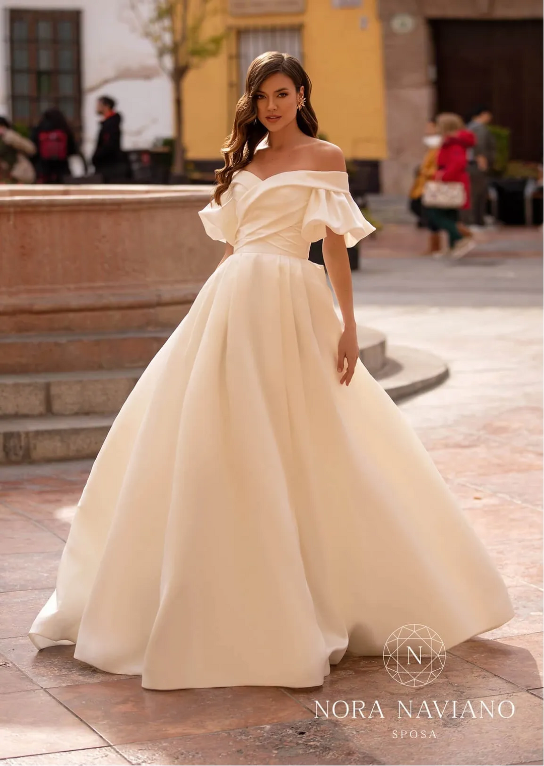 Woman in a white off-shoulder gown standing on a street with 'Nora Naviano' branding.