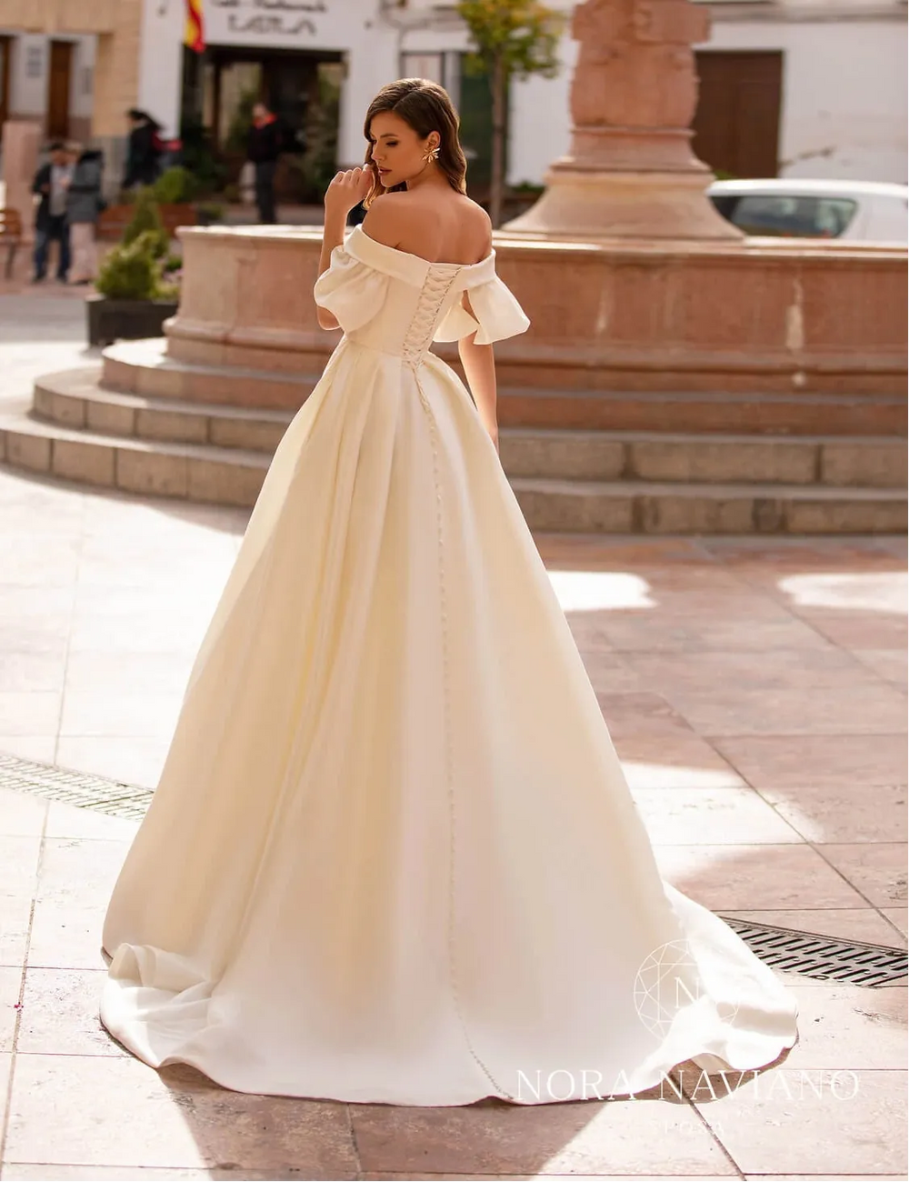 Woman in a white off-shoulder dress standing in front of a fountain in an urban setting.