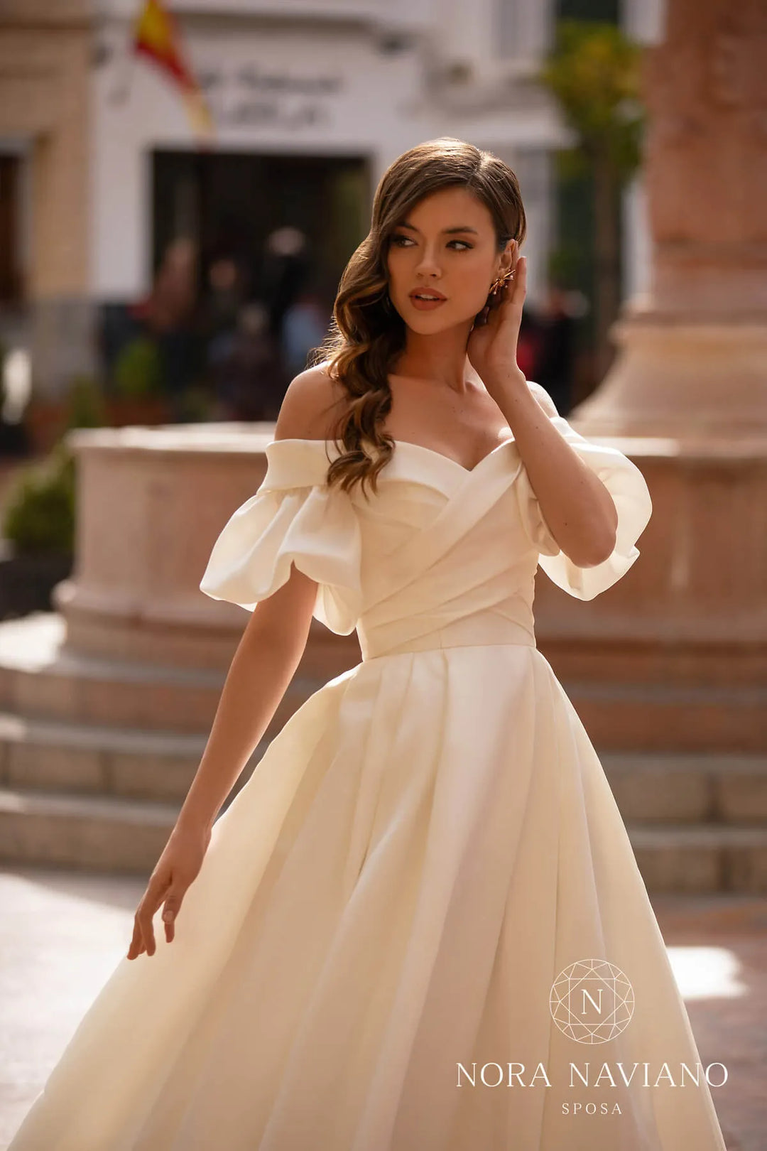 Woman in a white off-shoulder gown with 'Nora Naviano' branding in an outdoor setting.
