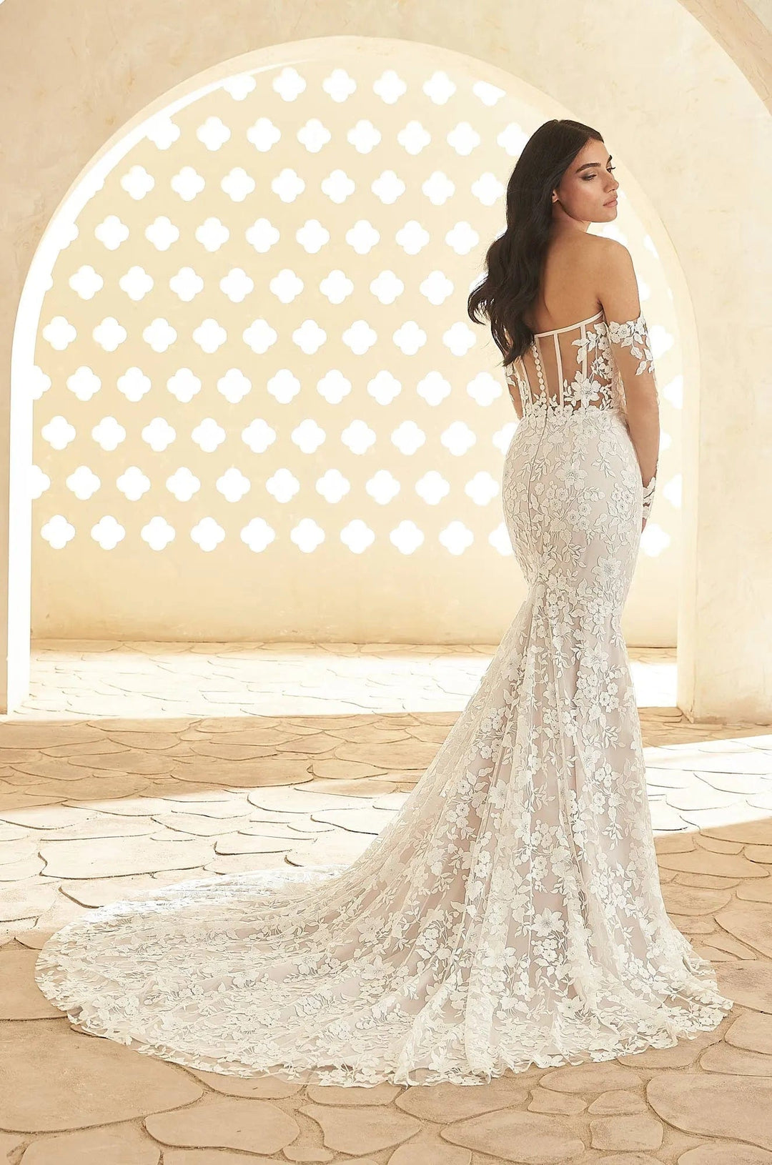 A woman wearing an off-the-shoulder mermaid-style wedding gown with intricate lace and bead embellishments, photographed from the back in a sunlit architectural setting with a stone archway.