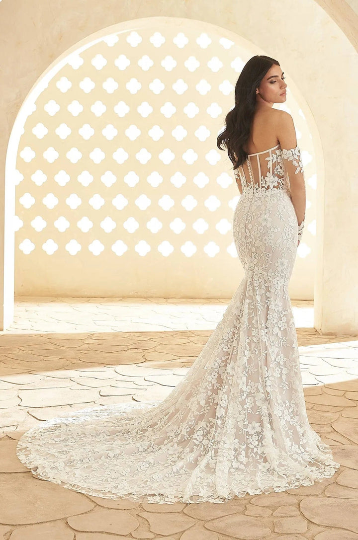 A woman wearing an off-the-shoulder mermaid-style wedding gown with intricate lace and bead embellishments, photographed from the back in a sunlit architectural setting with a stone archway.