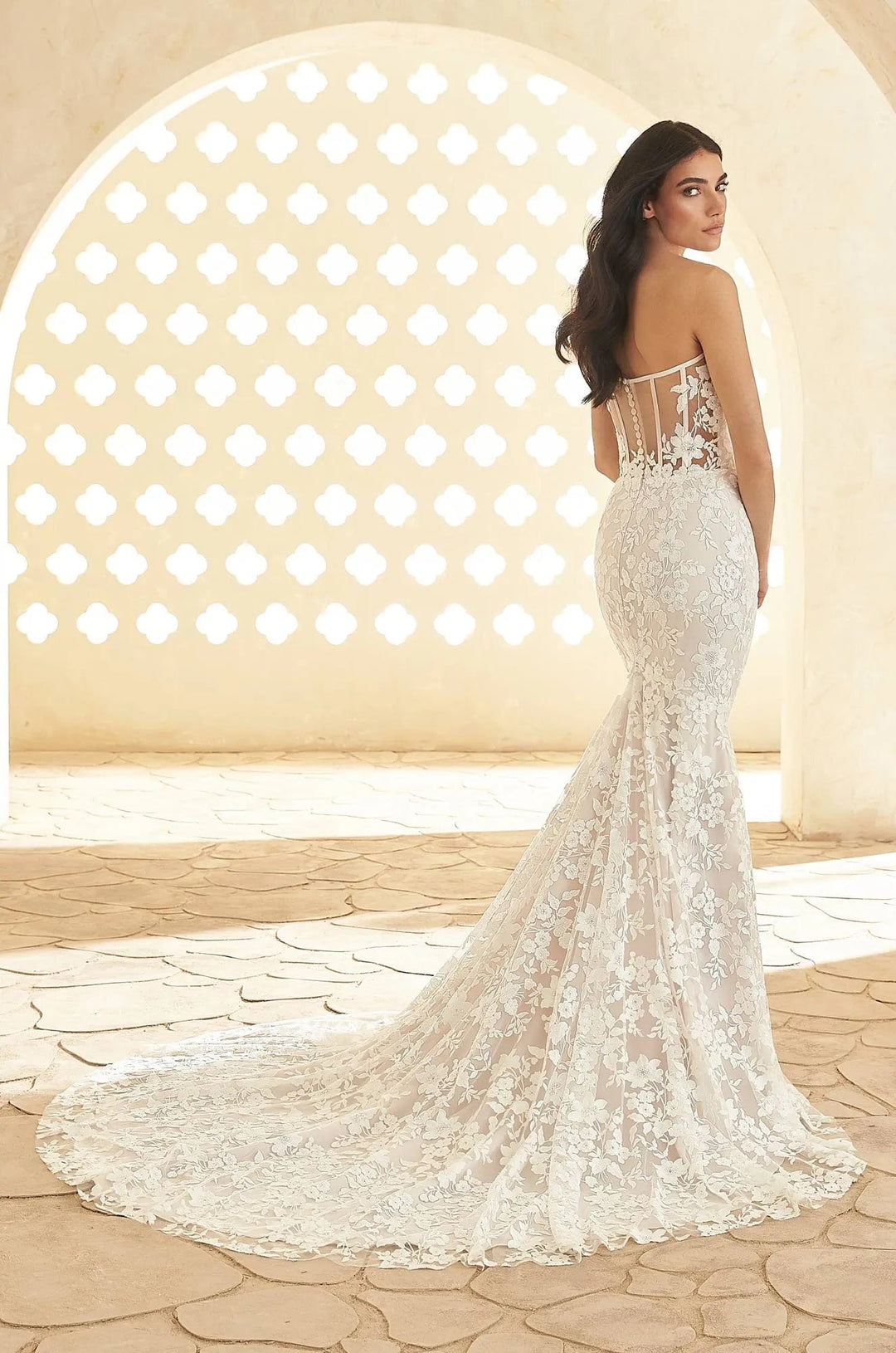 A woman wearing a strapless white lace mermaid wedding gown with a fitted bodice, sheer lace back, and long train, standing in a sunlit architectural setting with a stone arch and tiled floor.