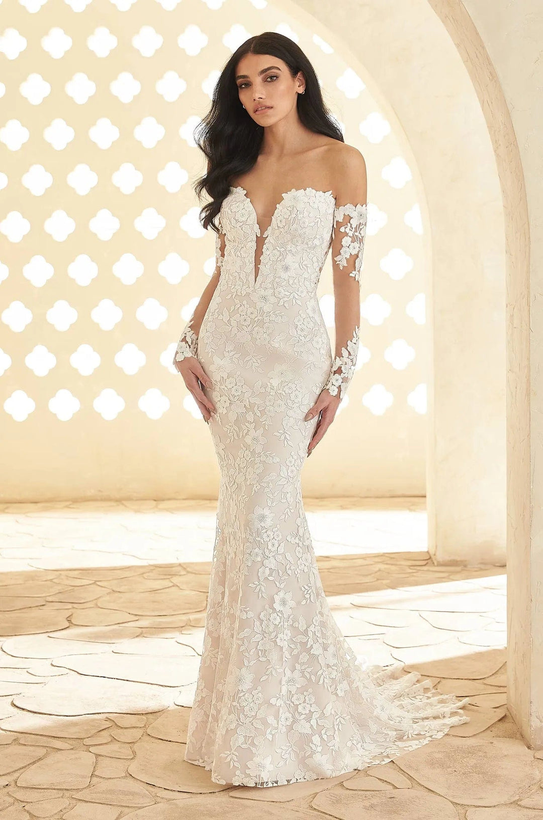 A woman modeling an off-the-shoulder white lace wedding gown with a deep V-neckline and long lace sleeves, standing in a sunlit architectural setting with arches and stone flooring.